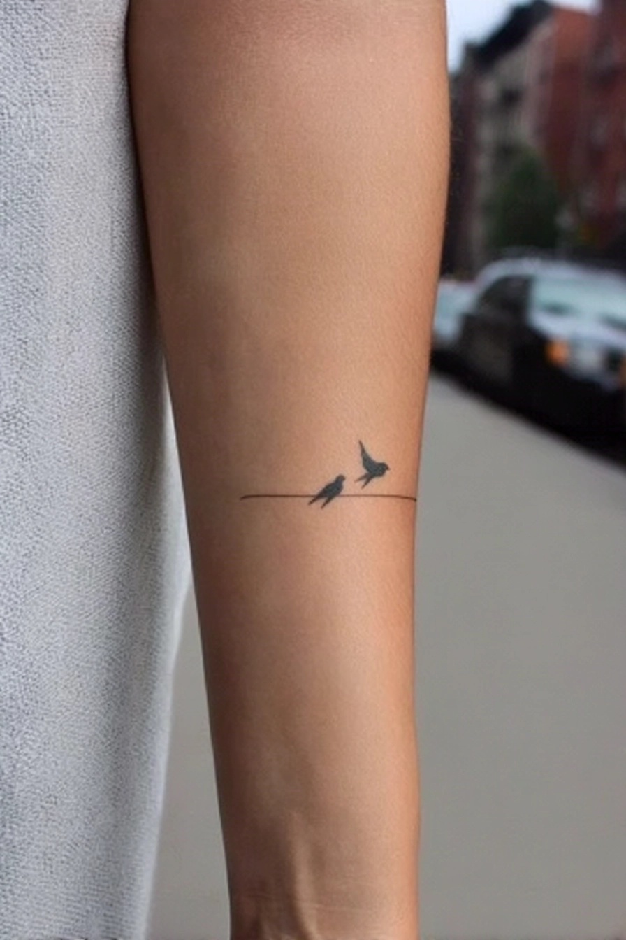 Minimalist tattoo of two birds sitting on a wire line on the forearm